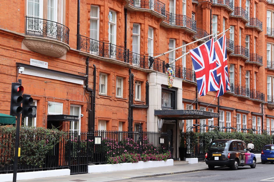 A multi-storey red brick building with ornate black wrought-iron balconies and white-framed windows, situated on a city street. A black metal fence lines the pavement in front of the building, with pink and purple flowering plants along its base. Two Union Jack flags are prominently displayed on flags attached to the building's facade. To the left, a pedestrian crossing signal displays a red light, while a traffic sign indicates the street name 'Davies Street W1.' A dark-colored taxi and a blue car are parked along the street, and a person is visible near the vehicle on the right. The scene captures an urban environment, likely during the daytime, with natural lighting illuminating the building's facade. This setting is indicative of central London, aligning with house removals and furniture transport activities, potentially involving Movers Mayfair or similar relocation services, as part of a professional home moving process in a busy city area.