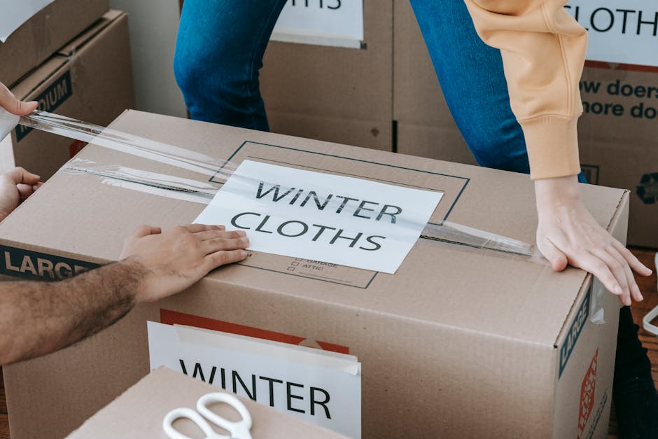 Close-up of a person positioning transparent packing tape on a large cardboard moving box labeled 'WINTER CLOTHS' in black text, with another person pressing down on the box to secure the tape. The scene shows multiple boxes stacked inside a property, with one box in the foreground and others partially visible in the background. The individuals are dressed in casual clothing, and the area appears to be part of a house or flat, ready for a home relocation or furniture transport. The boxes are made of corrugated cardboard, with printed labels indicating their contents, and a pair of scissors is partially visible on the box surface. The lighting is natural, and the setting reflects the packing process involved in a professional removals or moving service, such as those provided by Movers Mayfair.