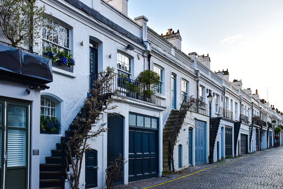 A row of white terraced houses on Brook Street in Mayfair, with each property featuring blue doors, window boxes, and external staircases leading from the pavement to the upper floors. The outside areas include small balconies with black metal railings, some with decorative plants, and neatly pruned trees alongside the staircases. The cobblestone street is visible in the foreground, with a slight incline, and the scene is illuminated by natural daylight. Movers Mayfair, a professional removals company, is involved in the home relocation process, which may include furniture transport, packing, and loading activities visible through the arrangement of boxes, furniture, and equipment such as trolleys or blankets near the entrances, ready for loading into moving vans.