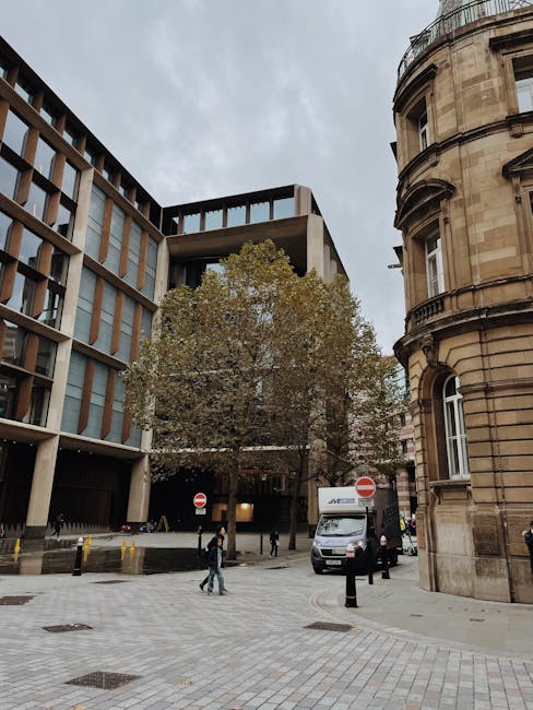 A wide street scene in Mayfair showing the exterior of an older, ornate sandstone building on the right with decorative window frames and rounded architectural features, contrasted by a modern glass and concrete office building on the left with large windows and a flat facade. In the centre, a large tree with sparse green and brown leaves partially obscures the view, indicating autumn. On the pavement, several pedestrians are walking, some carrying bags, while two movers from Movers Mayfair are seen loading a white moving van parked near the curb, which is partially obstructed by black bollards. The van’s rear doors are open, revealing packing boxes and furniture wrapped in plastic and blankets, prepared for home relocation. The surroundings include a clean, paved street with a few manhole covers, and overcast sky overhead, suggesting typical urban logistics involved in furniture transport and packing during house removals in central London.
