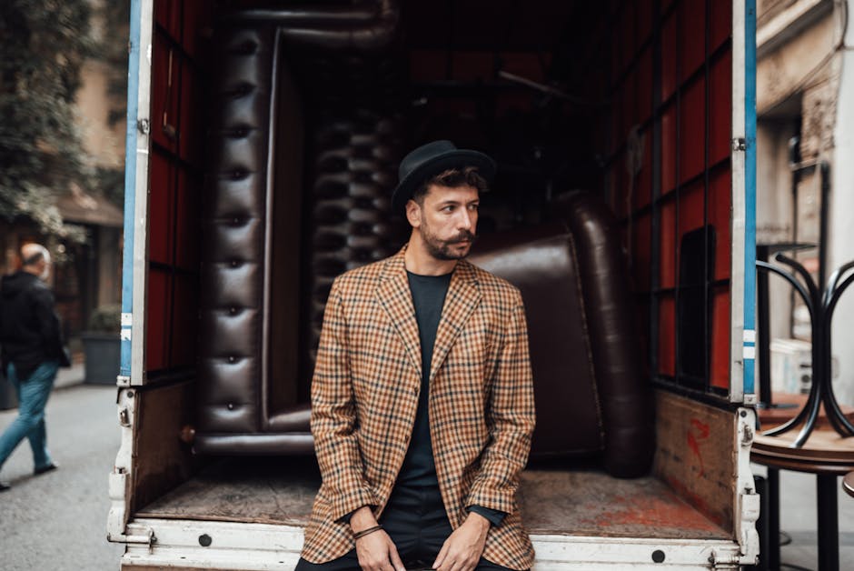 A man dressed in a black hat, checked blazer, and dark shirt stands inside the open back of a moving truck, which contains large, dark brown leather sofas and various packing materials such as cardboard boxes and protective blankets. The truck is parked on a street with a brick-paved surface, and nearby buildings with large windows are visible. In the background, a person is walking along the pavement, wearing casual clothing and a face mask. The scene depicts the loading process of a home relocation, with furniture and boxes being prepared for transport by a professional removals company, such as Movers Mayfair, in an urban environment consistent with house removals or furniture transport activities.