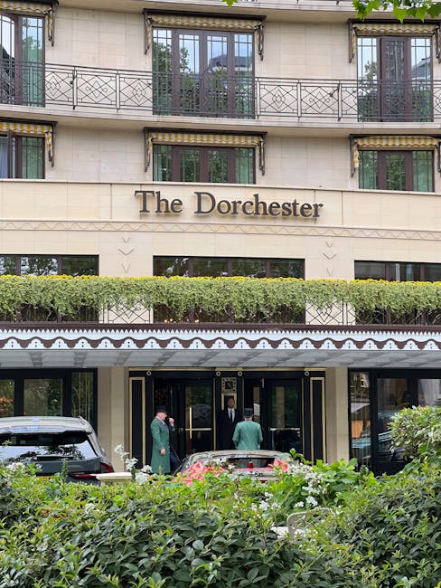 The exterior of The Dorchester hotel in Mayfair, London, showcasing its cream-colored stone façade with dark window frames and metal railings on the upper balconies. The entrance features black and gold detailing, including a canopy with decorative trim, and is flanked by well-maintained greenery, including neatly trimmed bushes. Several staff members dressed in formal dark suits and green uniforms are standing near the entrance, possibly preparing for a home relocation or assisting with a furniture transport process. A black vehicle is parked in front of the entrance, and the surrounding area includes lush shrubbery and pavement, indicating a busy and upscale hotel environment involved in logistics or moving services associated with house removals in Mayfair. Bright, natural light illuminates the scene, emphasizing the sophisticated architectural details of the building.