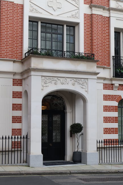 A row of red brick residential buildings with ornate architectural details, including bay windows, balconies with black wrought iron railings, and decorative white trim along the roofline, situated on