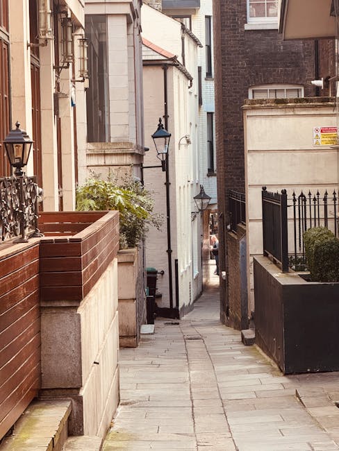 A narrow street in Mayfair with a paved walkway flanked by buildings on both sides, featuring brick and stone facades, some with metal fences and planters with greenery. On the left, a large wooden planter box with metal railings is mounted on a stone wall, while on the right, there is a black metal fence with decorative spikes and a small shrub. Tall, traditional street lamps are mounted on the walls, and the alleyway extends into the distance, showing glimpses of additional buildings and a downward slope. The scene suggests a typical London residential area, suitable for house removals or furniture transport, with visible elements of packing and moving logistics carried out by Movers Mayfair, evident from the environment prepared for a home relocation process.