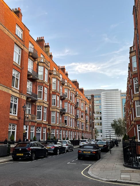 A view of a residential street lined with red-brick apartment buildings featuring multiple balconies, large windows, and decorative stonework. Several parked cars occupy the street alongside a sidewalk with a black metal fence and a small tree. In the background, a modern white office building contrasts with the older architecture, under a partly cloudy sky. The scene depicts an urban environment suitable for house removals and furniture transport services offered by Movers Mayfair, with visible elements of packing and loading processes typically involved in home relocation or moving logistics.