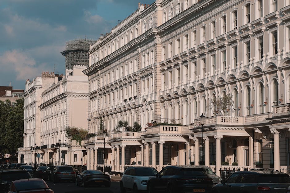 A brick apartment building on Mount Street features a black iron fence and a flowerbed with pink flowers along the sidewalk. Two Union Jack flags are displayed above the entrance, which has a dark canopy with the name 'Claridge's' visible. In front of the building, a black taxi and a blue car are parked on the street, with a traffic light showing red on the corner. Outside the building, a man is visible near a vehicle, possibly involved in a home relocation process. The scene suggests a busy area in Mayfair, where Movers Mayfair provides professional removals and furniture transport services, with equipment such as trolleys and packing materials potentially used in the loading process inside or around the property.