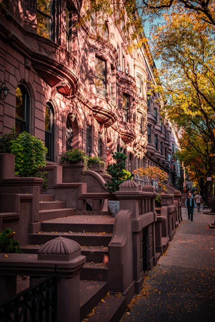 The image shows a row of brownstone residential buildings with ornate architectural details, such as decorative window surrounds and small balconies, situated along a tree-lined street in Mayfair. The buildings are accessed via steps leading up from the sidewalk, which is lined with a black wrought iron fence and small planters filled with green foliage. Autumn leaves are scattered on the pavement, and the trees above have leaves in shades of orange and yellow, indicating a fall setting. In the foreground, a person dressed in dark clothing is walking along the sidewalk, while other pedestrians are visible further along the street. The scene is captured during daylight with dappled sunlight filtering through the branches. The setting suggests a typical residential area suitable for house removals and furniture transport, with a focus on the urban environment and the process of moving home. The image subtly supports the theme of packing and moving logistics, as it depicts a typical street scene where professional movers like Movers Mayfair might operate during a home relocation involving bulky furniture or antiques.