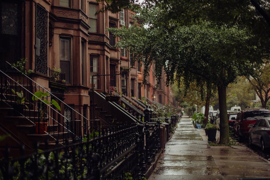 A row of brownstone residential buildings on a wet city street during daytime, with exterior stairs leading to front doors decorated with potted plants and flowers. The sidewalk is lined with green leafy trees and some planters, with a few parked cars along the curb. In the foreground, a professional moving team from Movers Mayfair is visible, carefully loading a large, wrapped antique wooden wardrobe onto a moving van parked nearby. The team uses protective blankets and straps to secure the furniture, and the scene captures the home relocation and furniture transport process on a damp day, emphasizing careful packing and loading for house removals in Mayfair.