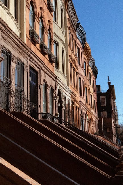 The image shows a row of brownstone residential buildings with ornate architectural details, such as decorative window surrounds and small balconies, situated along a tree-lined street in Mayfair. The buildings are accessed via steps leading up from the sidewalk, which is lined with a black wrought iron fence and small planters filled with green foliage. Autumn leaves are scattered on the pavement, and the trees above have leaves in shades of orange and yellow, indicating a fall setting. In the foreground, a person dressed in dark clothing is walking along the sidewalk, while other pedestrians are visible further along the street. The scene is captured during daylight with dappled sunlight filtering through the branches. The setting suggests a typical residential area suitable for house removals and furniture transport, with a focus on the urban environment and the process of moving home. The image subtly supports the theme of packing and moving logistics, as it depicts a typical street scene where professional movers like Movers Mayfair might operate during a home relocation involving bulky furniture or antiques.