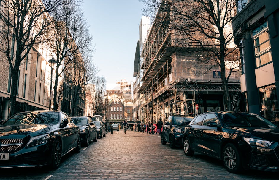 A street scene in Mayfair with several dark-colored cars parked along the cobblestone pavement, which is lined with leafless trees and modern office buildings. One building on the right has scaffolding and protective netting indicating ongoing construction or refurbishment. The pavement is occupied by people, some of whom appear to be part of a home relocation or moving process, possibly waiting or preparing to load furniture or boxes into a moving van. The area is well-lit by natural daylight, with a clear sky overhead. The presence of construction materials and parked vehicles suggests that this location may be affected by parking suspensions and fines, relevant to house removals in Mayfair, as managed by Movers Mayfair. The scene captures a typical urban environment where loading and unloading activities take place during a move, with no visible moving equipment or furniture currently being transported but indicating the logistical context of a move or furniture transport operation within the busy city street.