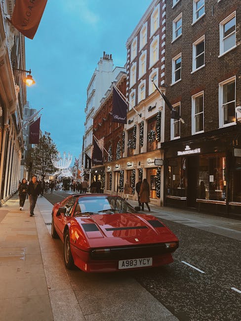 A red vintage sports car is parked on a busy city street in Mayfair, with pedestrians walking on the pavement and storefronts lining both sides of the road. The car's sleek design and low profile are clearly visible, along with its distinctive front hood and license plate. In the background, multi-storey buildings feature traditional brick facades, large windows, and hanging flags, some of which are part of nearby shops and restaurants. Street lighting illuminates the scene as the sky transitions from daylight to early evening. This setting depicts a typical urban environment suitable for home relocation or furniture transport by professional removals services such as Movers Mayfair, who coordinate loading and unloading processes in such busy commercial and residential areas.