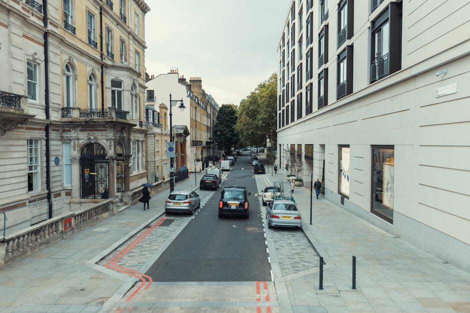 A street scene in Mayfair with several dark-colored cars parked along the cobblestone pavement, which is lined with leafless trees and modern office buildings. One building on the right has scaffolding and protective netting indicating ongoing construction or refurbishment. The pavement is occupied by people, some of whom appear to be part of a home relocation or moving process, possibly waiting or preparing to load furniture or boxes into a moving van. The area is well-lit by natural daylight, with a clear sky overhead. The presence of construction materials and parked vehicles suggests that this location may be affected by parking suspensions and fines, relevant to house removals in Mayfair, as managed by Movers Mayfair. The scene captures a typical urban environment where loading and unloading activities take place during a move, with no visible moving equipment or furniture currently being transported but indicating the logistical context of a move or furniture transport operation within the busy city street.