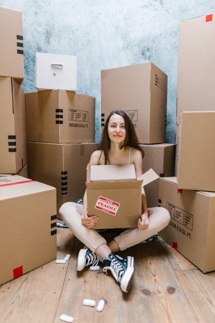 A young woman with long dark hair, sitting on a wooden floor inside a room with a textured blue wall, surrounded by numerous cardboard boxes of various sizes, some sealed with packing tape and others open. She is holding a medium-sized cardboard box with a red 'Fragile' sticker on the front, indicating careful handling during a home relocation process. The boxes are stacked against the wall, and some are positioned at different heights, suggesting an active packing or unpacking phase typical of furniture transport or packing and moving services. Visible at the feet are small white packing peanuts scattered on the floor, and behind her is a doorway that offers partial view into the space where the boxes are stored or prepared for transport. The scene reflects a typical moving environment managed by Movers Mayfair, with a focus on packing materials, logistical organisation, and the movement of household items inside a property.