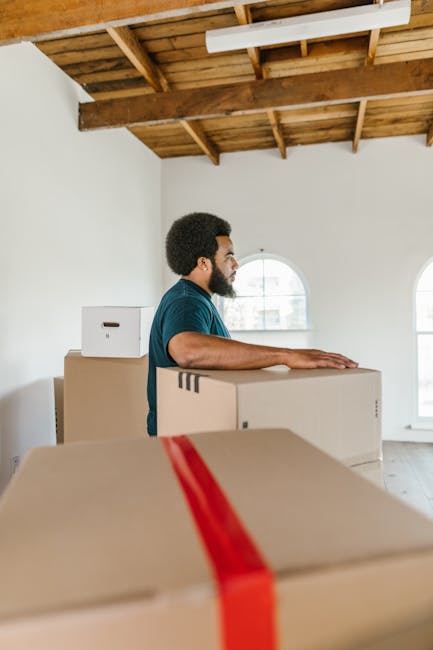 A man with dark curly hair and a beard, dressed in a blue t-shirt, is inside a room with white walls and a wooden ceiling with exposed beams. He is standing behind several large cardboard moving boxes stacked at waist height, with one box in the foreground wrapped with red packing tape. The room features two arched windows allowing natural light to illuminate the space. The scene depicts the process of home relocation, with the man preparing boxes for packing or transport, indicative of furniture transport and packing and moving operations. This image exemplifies a professional removal process, likely carried out by movers such as Movers Mayfair, in a typical residential setting during a same-day emergency move or planned home removal, as the boxes are ready for loading onto a moving vehicle.