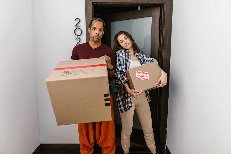 A young woman with long dark hair, sitting on a wooden floor inside a room with a textured blue wall, surrounded by numerous cardboard boxes of various sizes, some sealed with packing tape and others open. She is holding a medium-sized cardboard box with a red 'Fragile' sticker on the front, indicating careful handling during a home relocation process. The boxes are stacked against the wall, and some are positioned at different heights, suggesting an active packing or unpacking phase typical of furniture transport or packing and moving services. Visible at the feet are small white packing peanuts scattered on the floor, and behind her is a doorway that offers partial view into the space where the boxes are stored or prepared for transport. The scene reflects a typical moving environment managed by Movers Mayfair, with a focus on packing materials, logistical organisation, and the movement of household items inside a property.