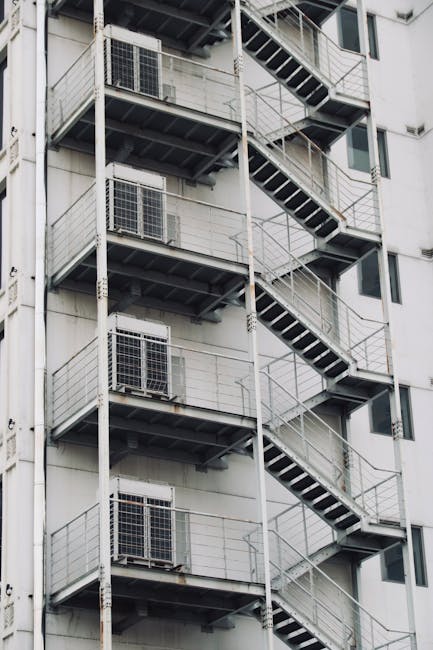 A tall, exterior metal fire escape staircase attached to the side of a white residential building with multiple floors. Each level features a small landing with metal railings and steps leading upward, supported by vertical metal poles. The building has several windows, some with visible air conditioning units. The staircase is black with non-slip treads, and the structure appears weathered with some rust visible. The scene is well-lit with natural daylight, highlighting the industrial design of the staircase and the building's facade, which is part of the context for home relocation or furniture transport through stair access rules for Mayfair flats, as handled by Movers Mayfair.