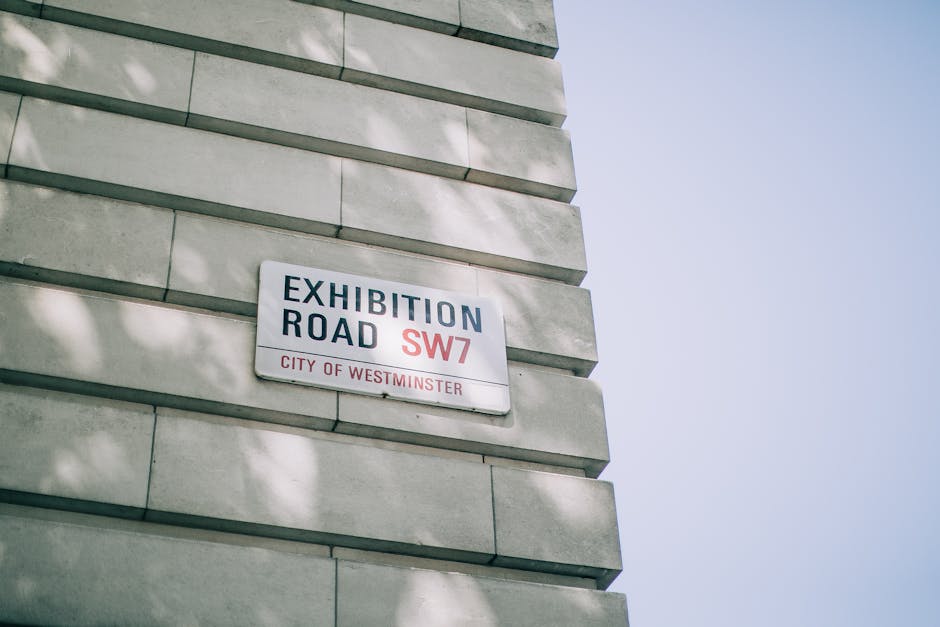 A close-up view of a street sign mounted on a light-colored stone wall, displaying 'Exhibition Road SW7' and indicating it is in the City of Westminster. The sign is rectangular, with a white background, black and red text, and is attached to the wall beneath overhanging tree shadows. In the background, a clear sky is visible, suggesting daytime. The photograph captures part of the building's facade, which consists of horizontally aligned stone blocks with subtle shadows indicating the layered construction. The setting relates to the London area, where house removals and moving services by Movers Mayfair often operate, emphasizing urban relocation logistics and an environment suitable for furniture transport and home moving procedures.