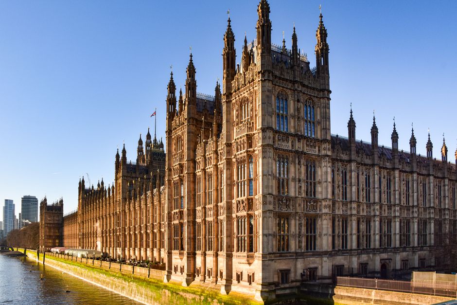 A wide-angle view of the Palace of Westminster illuminated by sunlight, showcasing the detailed Gothic architecture with tall spires and ornate stonework, situated along the River Thames in London. The reflection of the building is visible in the water, with a clear blue sky overhead. In the foreground, the riverbank is visible with a grassy edge and part of a paved walkway, while modern high-rise buildings are faintly seen in the background. The image captures the iconic landmarks typical of central London, relevant to house removals and moving services such as those provided by Movers Mayfair, illustrating the environment related to property relocation and transportation within the city.