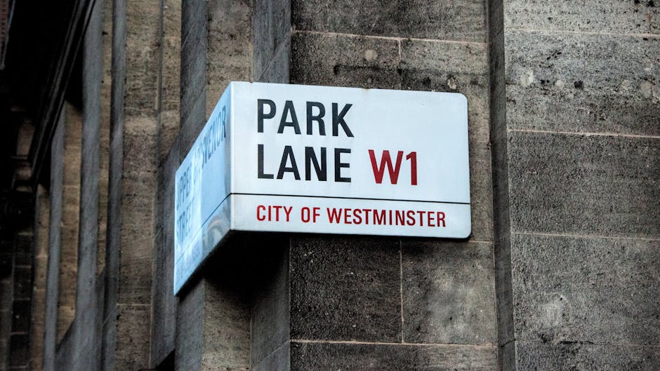 A close-up view of a street sign mounted on a light-colored stone wall, displaying 'Exhibition Road SW7' and indicating it is in the City of Westminster. The sign is rectangular, with a white background, black and red text, and is attached to the wall beneath overhanging tree shadows. In the background, a clear sky is visible, suggesting daytime. The photograph captures part of the building's facade, which consists of horizontally aligned stone blocks with subtle shadows indicating the layered construction. The setting relates to the London area, where house removals and moving services by Movers Mayfair often operate, emphasizing urban relocation logistics and an environment suitable for furniture transport and home moving procedures.